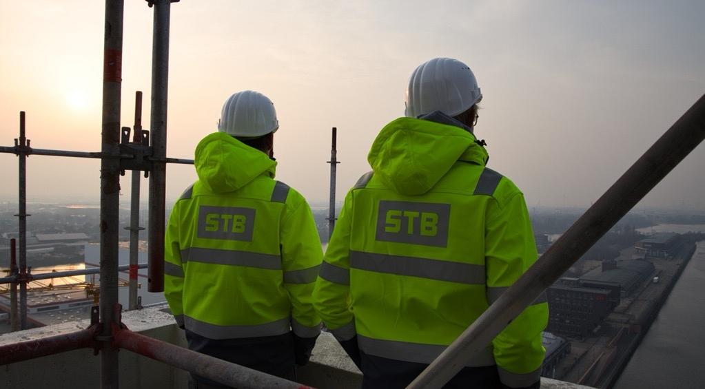 Zwei Kollegen in gelben Warnjacken mit STB-Logo stehen mit dem Rücken zur Kamera auf eine Baustelle und schauen in den Sonnenaufgang.
