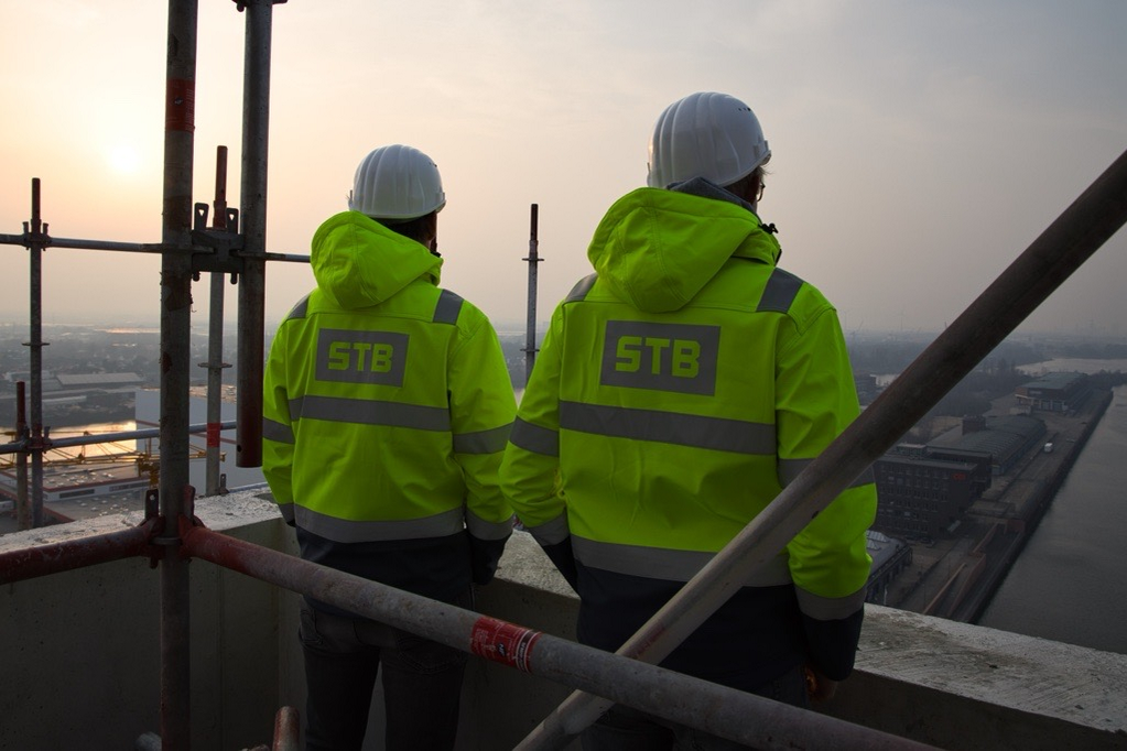 Zwei Kollegen in gelben Warnjacken mit STB-Logo stehen mit dem Rücken zur Kamera auf eine Baustelle und schauen in den Sonnenaufgang.