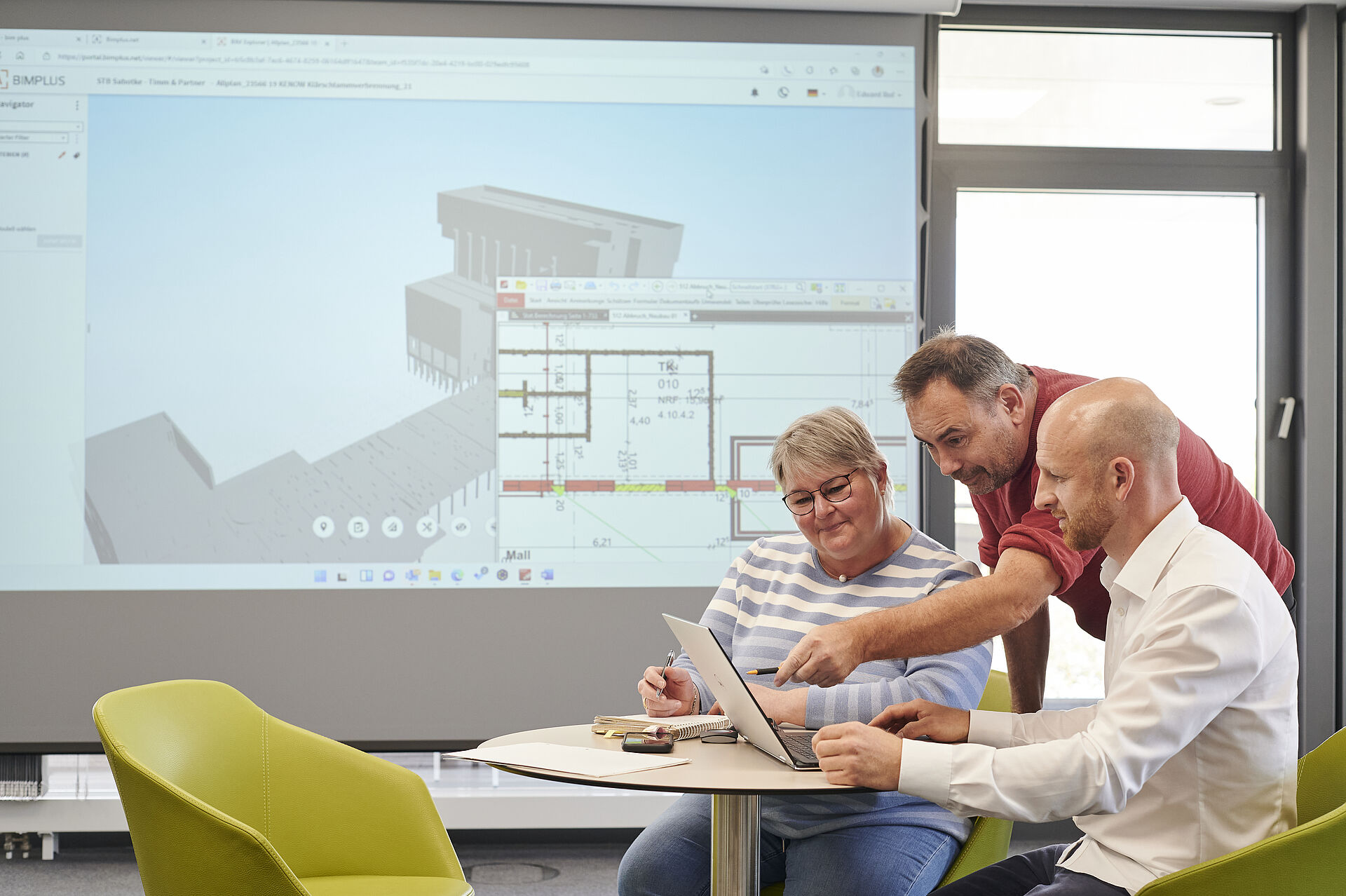 Drei Kolleg*innen sitzen an einem Tisch auf dem ein Laptop steht. Im Hintergrund ist ein Beamer mit einem CAD-Modell in 3D zu sehen.